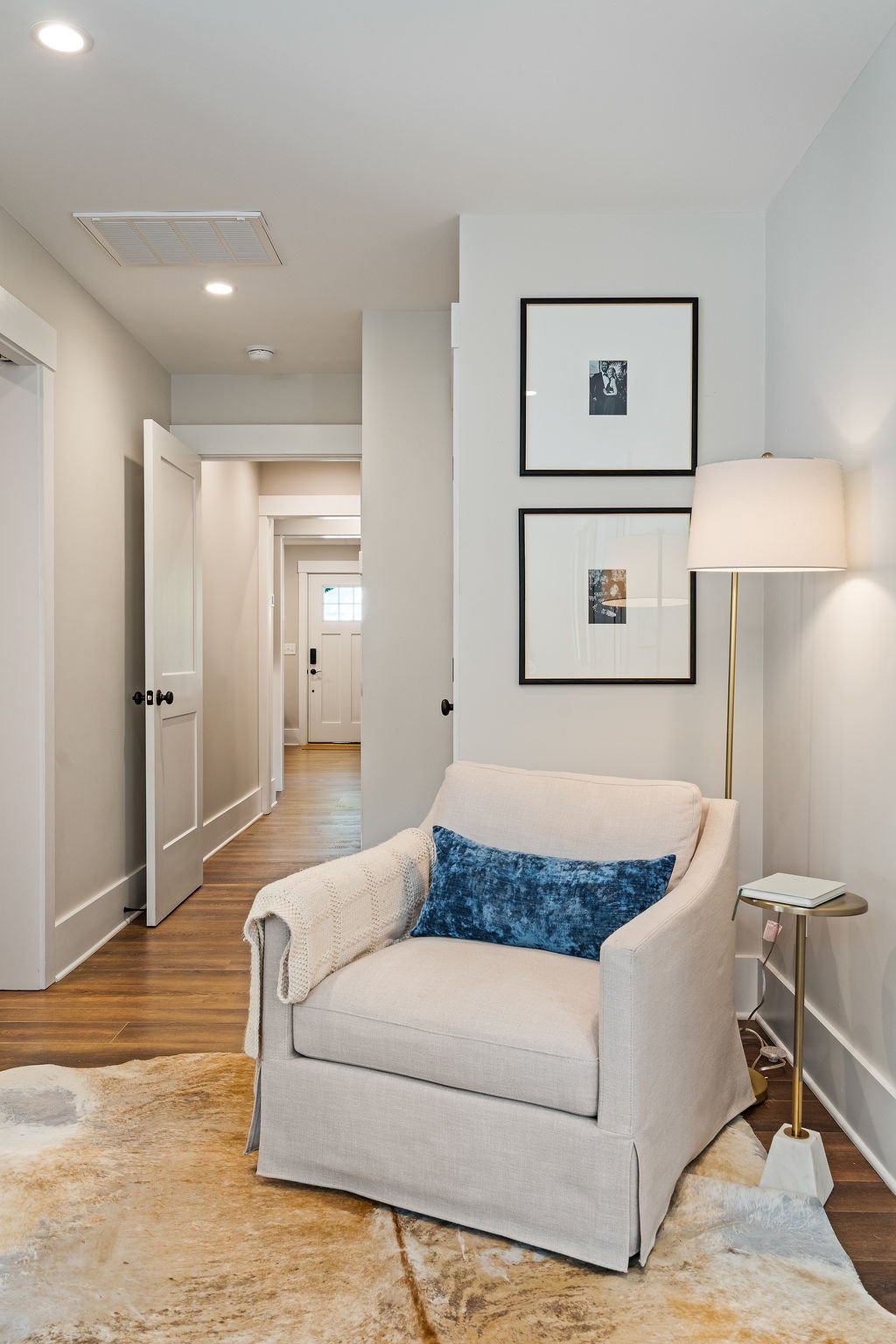 Reading nook with a white chair, blue pillow, side table, floor lamp, two framed pictures hanging above it, white walls and a hallway leading away.