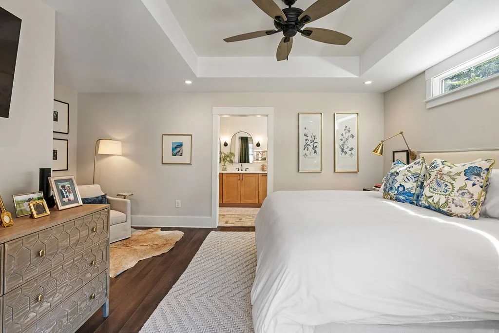 Interior design shot of bedroom showing dresser with photos, bed with white comforter on white rug, colorful throw pillows, several pieces of art on the walls, reading nook and doorway to the bathroom.