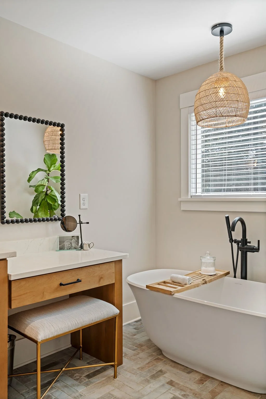 Corner of bathroom showing freestanding tub, tub shelf, large window, wicket pendant lighting and desk, stool and mirror next to it.