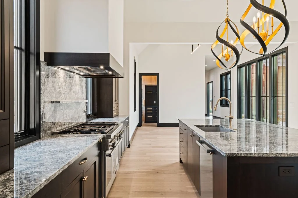 Modern kitchen with granite countertops, gas stove, sink and hanging gold lights.