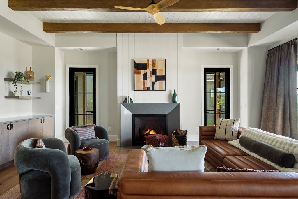 Modern living room with wooden beams, leather couch, fireplace, abstract art above the concrete mantle and glass doors on either side.