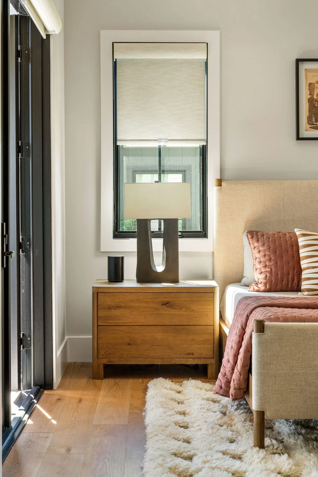 Bedside table with modern lamp, door frame on the left and the bed with art above it and red throw pillows on it.