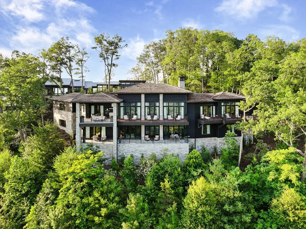 Aerial architectural image of a multi-level custom mountain home with stone walls, large windows, outdoor terraces and wooded surroundings.