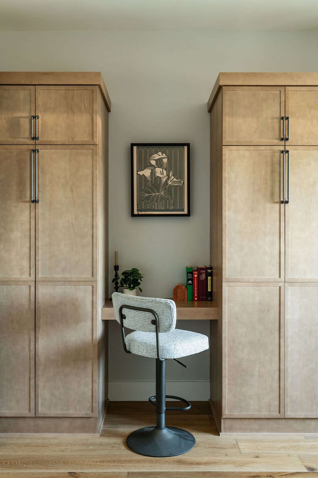 Bedroom desk set between two tall cabinets with black and gold art above it, books and a plant on it, and a grey chair in front.
