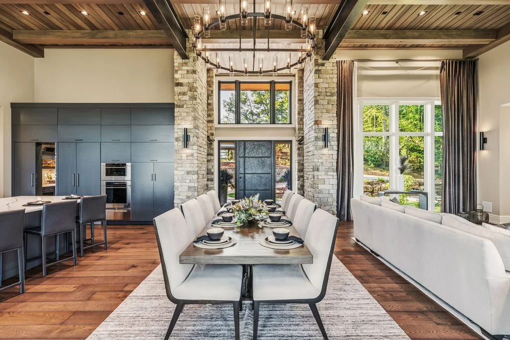 Open-concept home, showing kitchen on the left, dining room with chandelier in the middle, and living room on the right. The front door is past the table and there is a wood ceiling with beams.