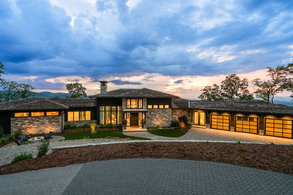 Luxury home at twilight under a cloudy sky with stone walls, dark siding, illuminated windows, a fire pit and three-car garage.