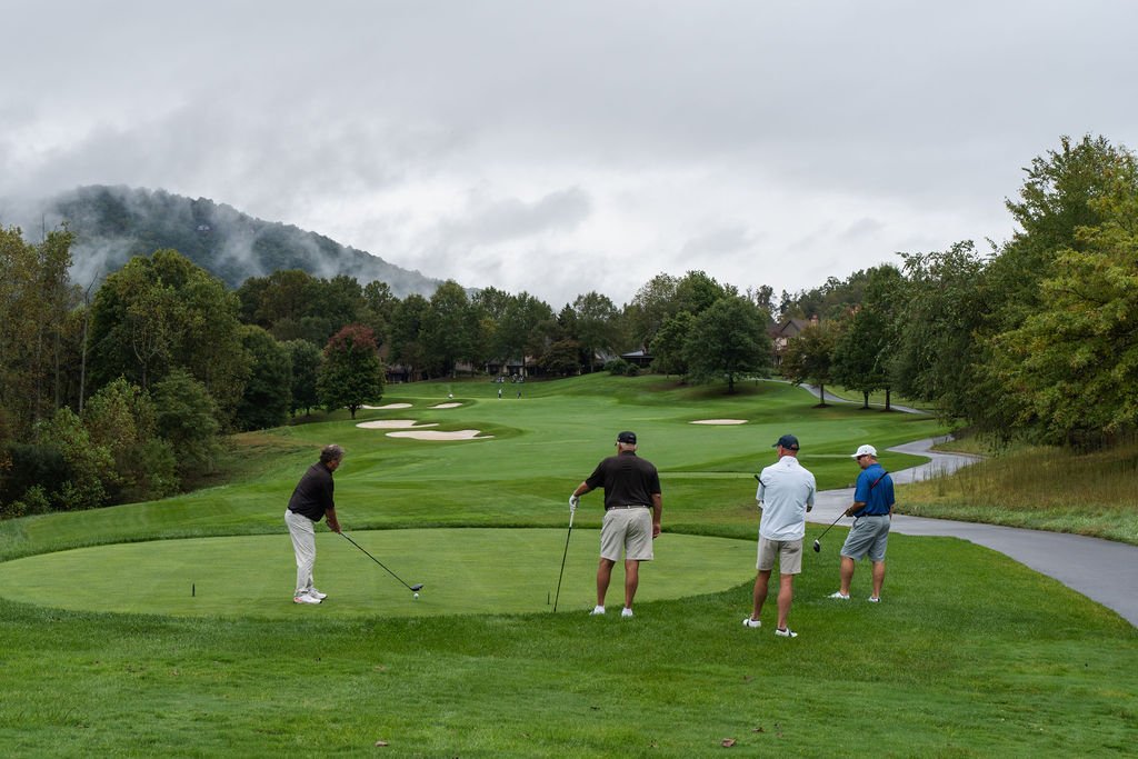 Group of three men watching another hit a golf ball in a tee box on a lush green course on a foggy day.
