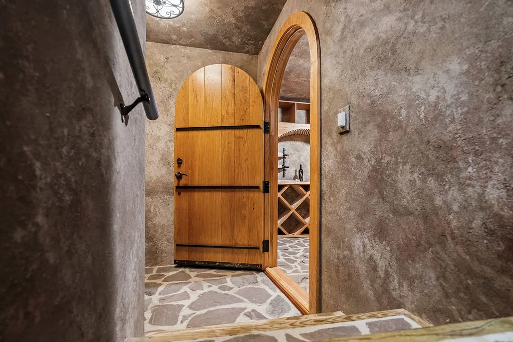Rustic custom wine cellar entrance with an arched oak door, textured plaster walls, stone flooring and built-in bottle storage.