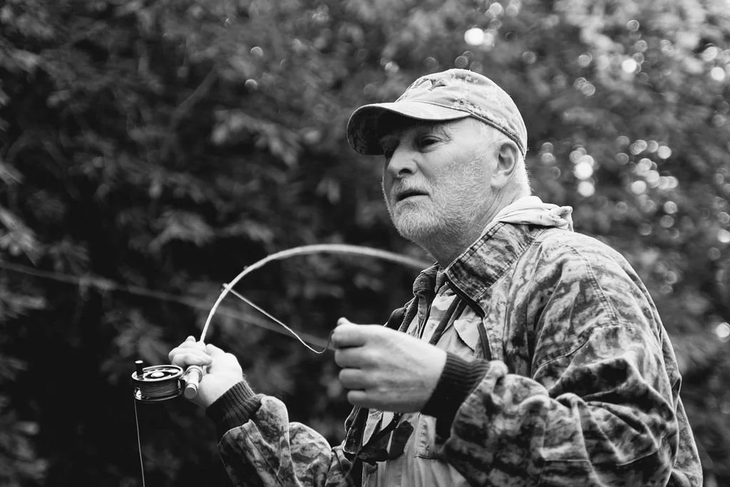 Black-and-white portrait of an elderly man wearing a hat and camo jacket while fly fishing.