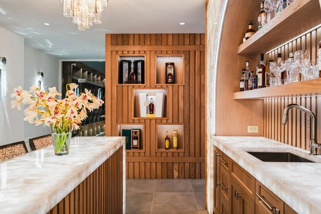Modern home bar with lighted marble countertops, custom wood walls and shelving and a variety of liquor bottles.