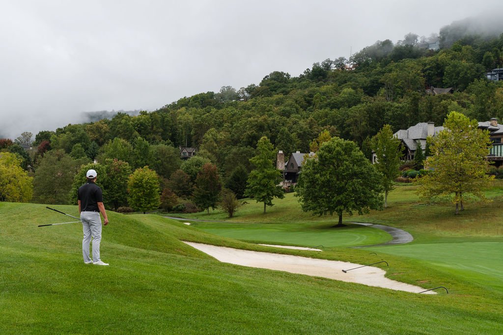 Man in golf attire standing on a hill in the fairway overlooking the course with houses set in the heavily wooded tree line.