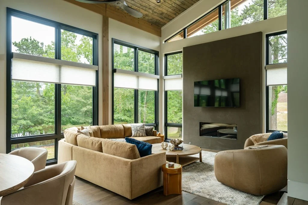 Modern living room with floor-to-ceiling windows, wooden ceiling, tan couch with blue pillows, two leather chairs and a coffee table facing the fireplace/tv.