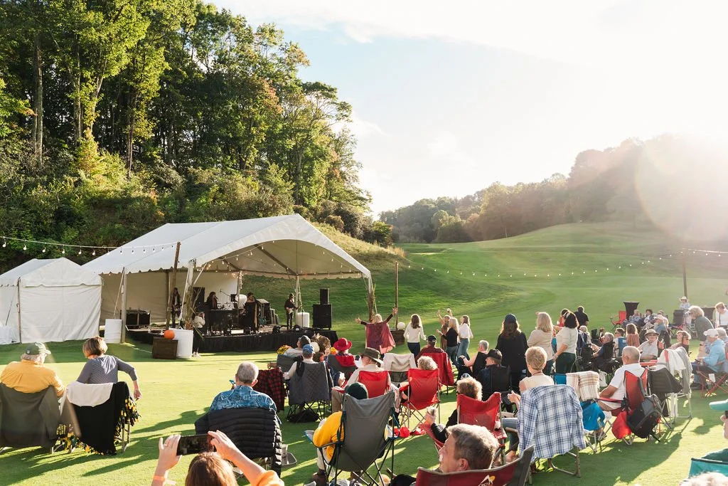 Concert on a golf course surrounded by trees with the band playing under a white tent and the crowd sitting in lawn chairs.