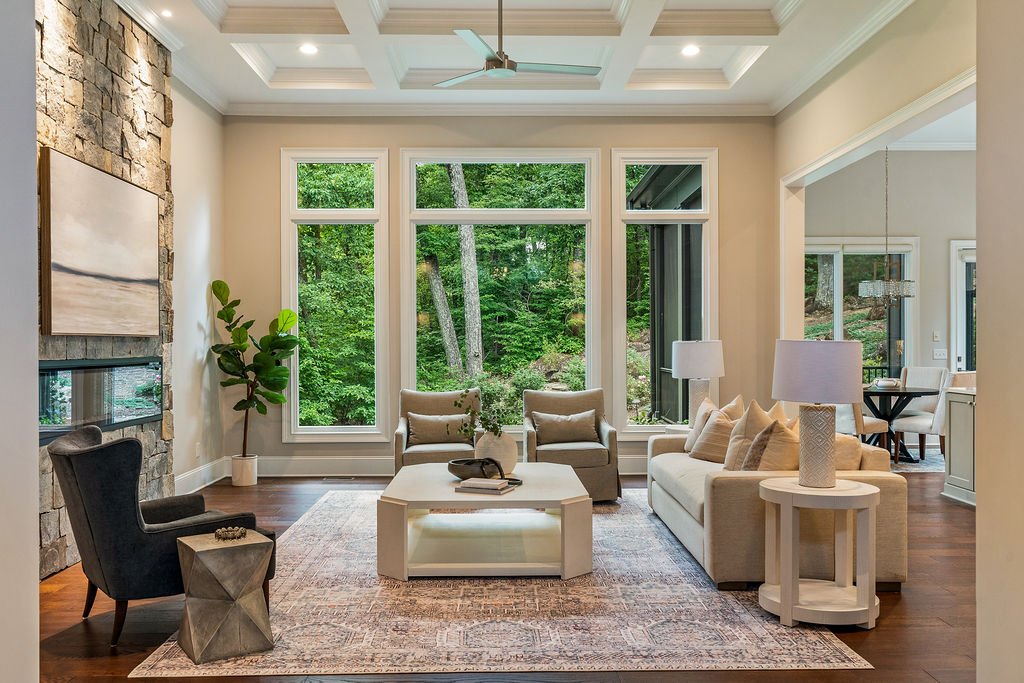 Living room with a stone fireplace, coffered ceiling, neutral furnishings and expansive windows facing the woods.