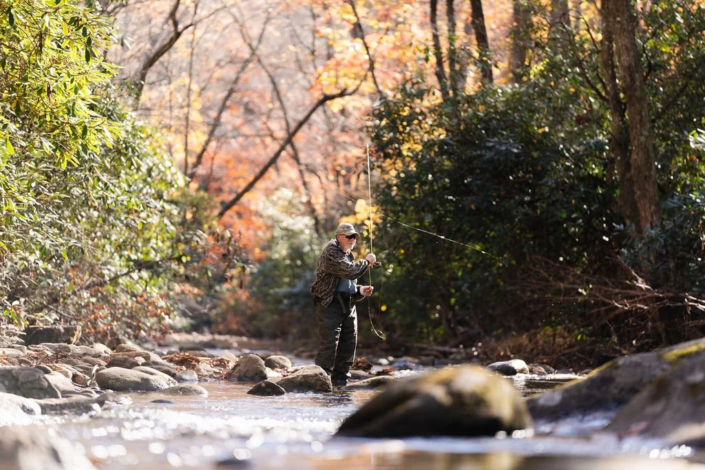 Man standing in the middle of a shallow river among rocks and fishing with fall colored trees in the background.