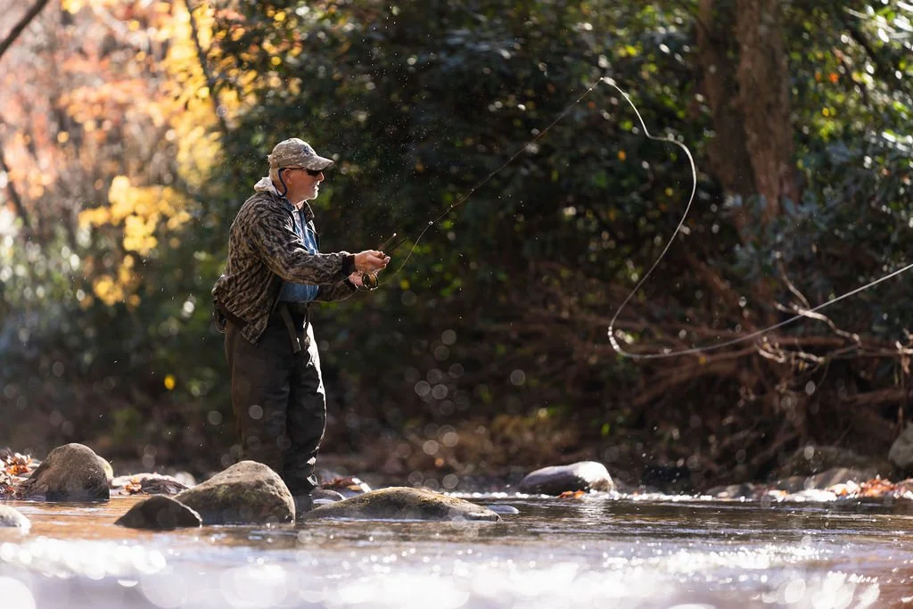 Man fly fishing in shallow river surrounded by vibrant fall-colored trees.