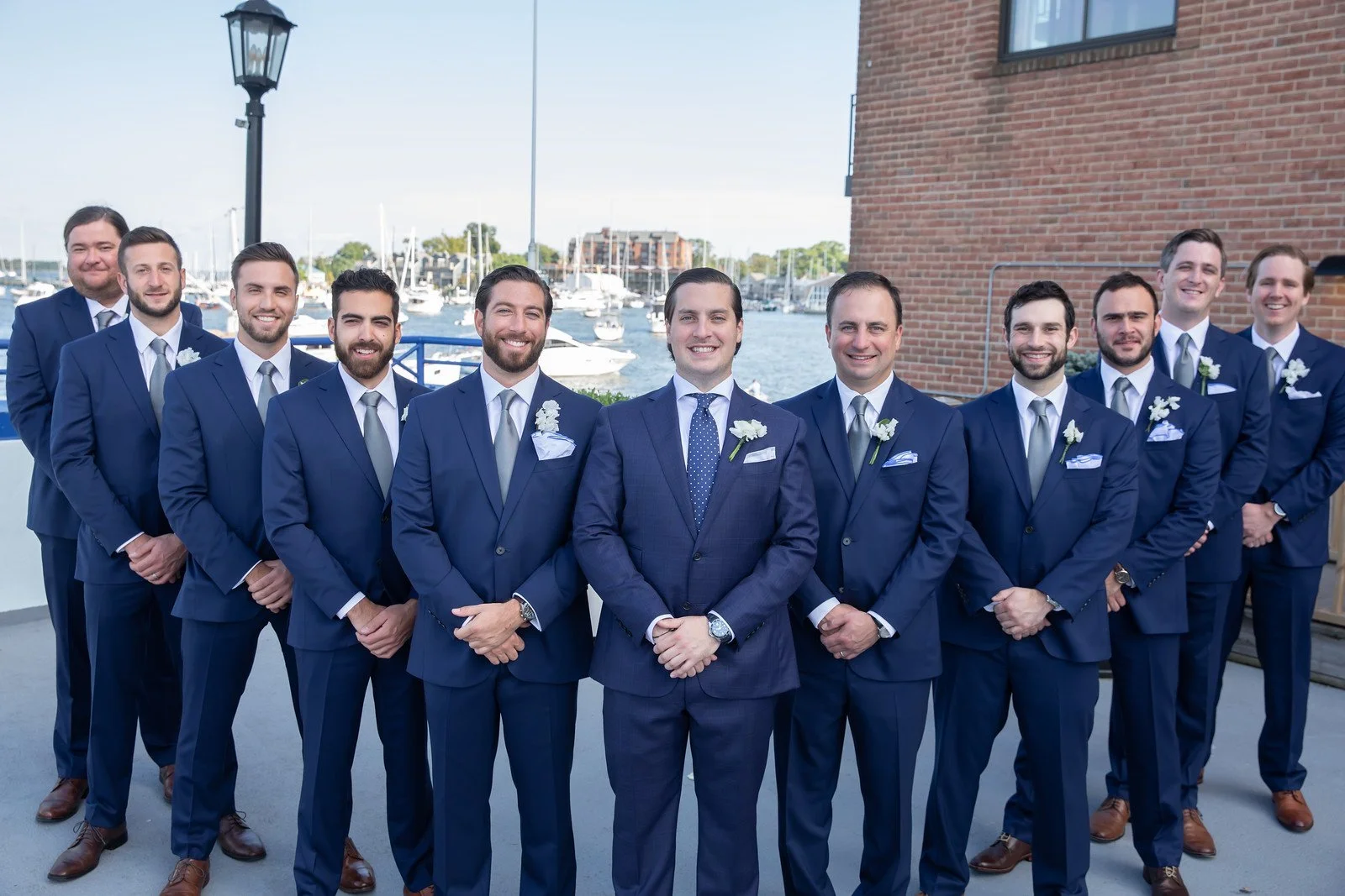 A group of eleven men dressed in matching navy suits, white shirts, and ties, standing outdoors near a waterfront with boats, a brick building, and a lamp post in the background.