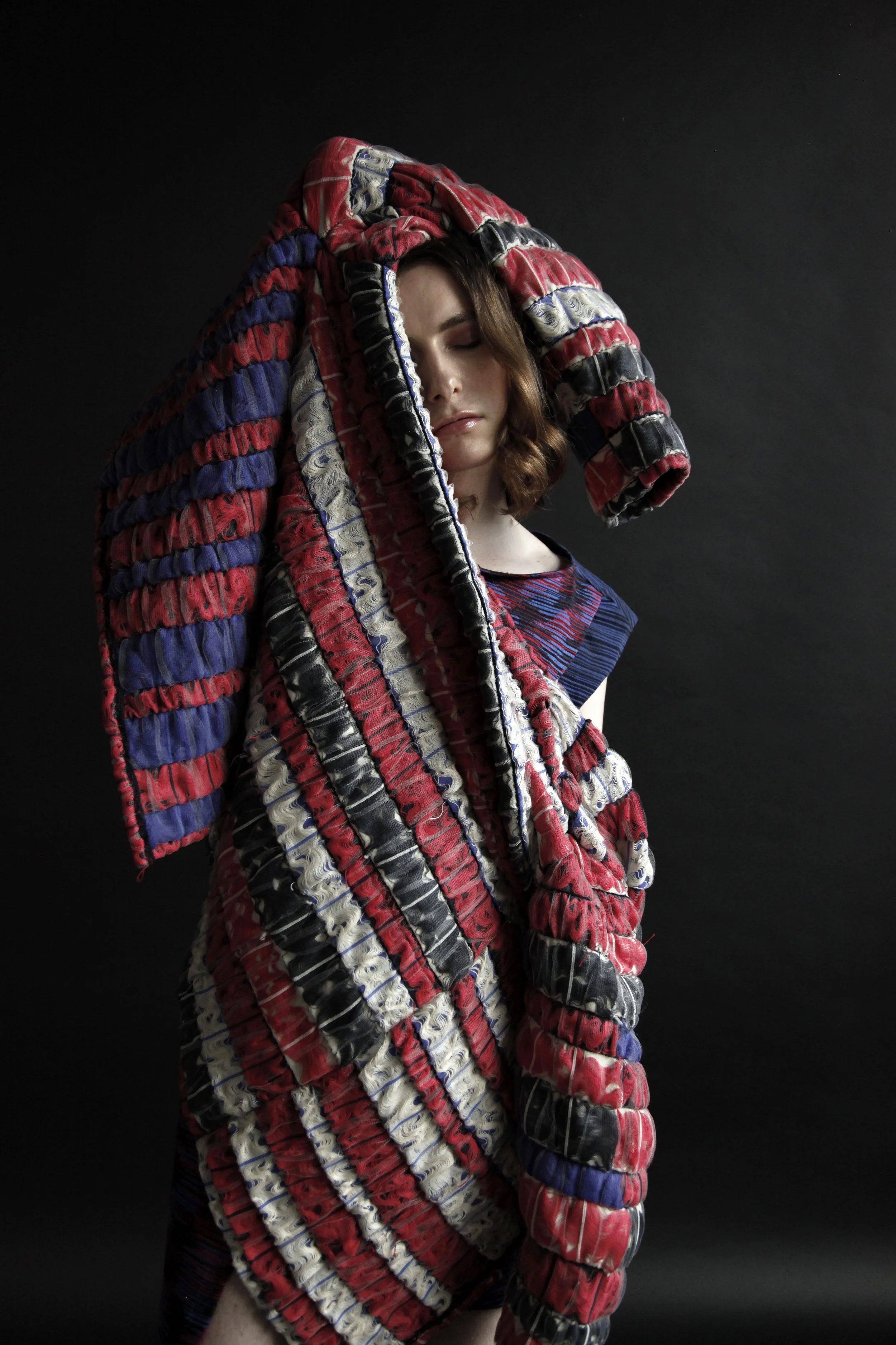 A woman with closed eyes poses against a dark background, wrapped in a large, colorful, quilted fabric with red, black, white, and blue patches.