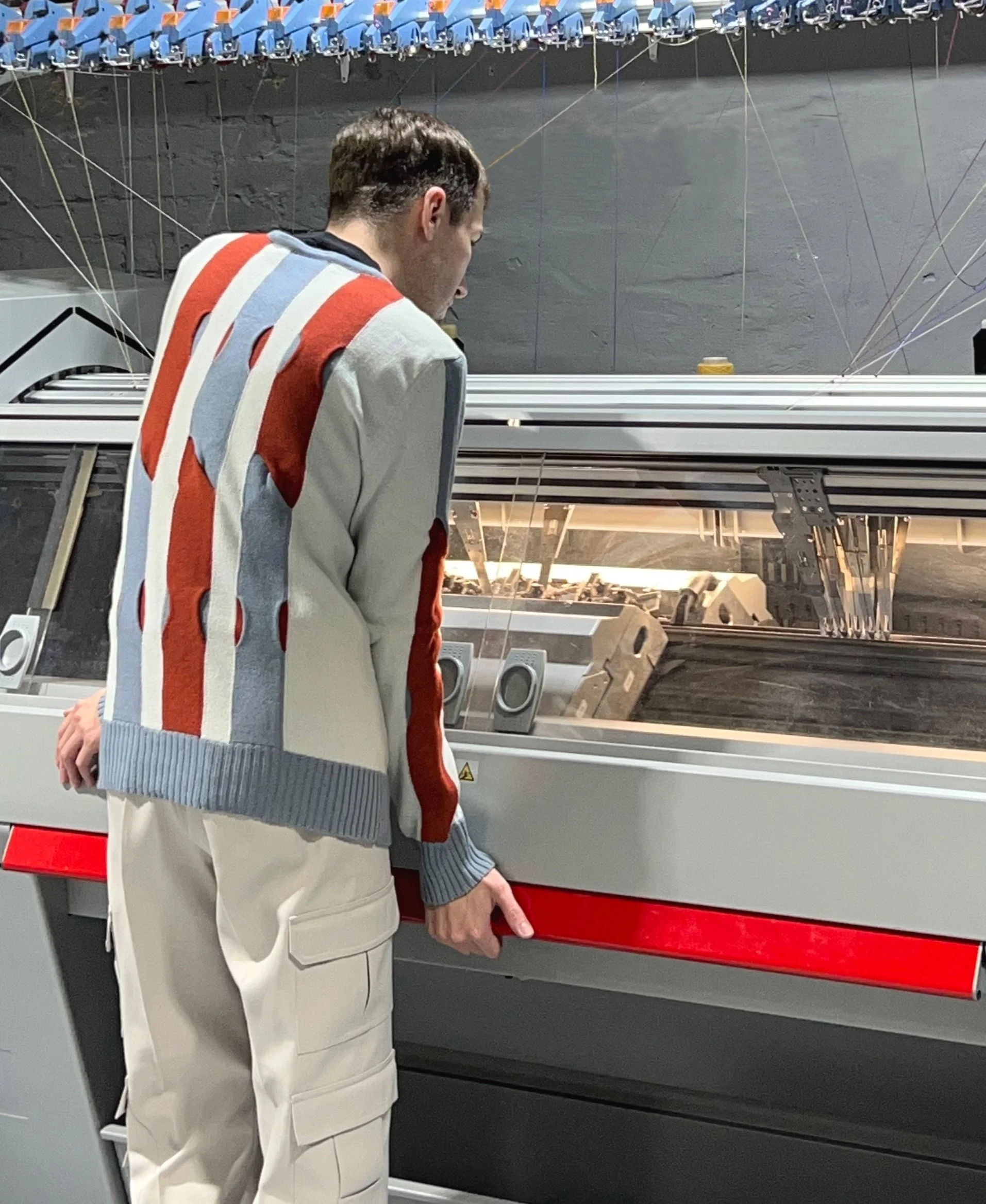 A young man wearing a multicolored striped sweater stands in front of a large industrial machine, observing it closely indoors.