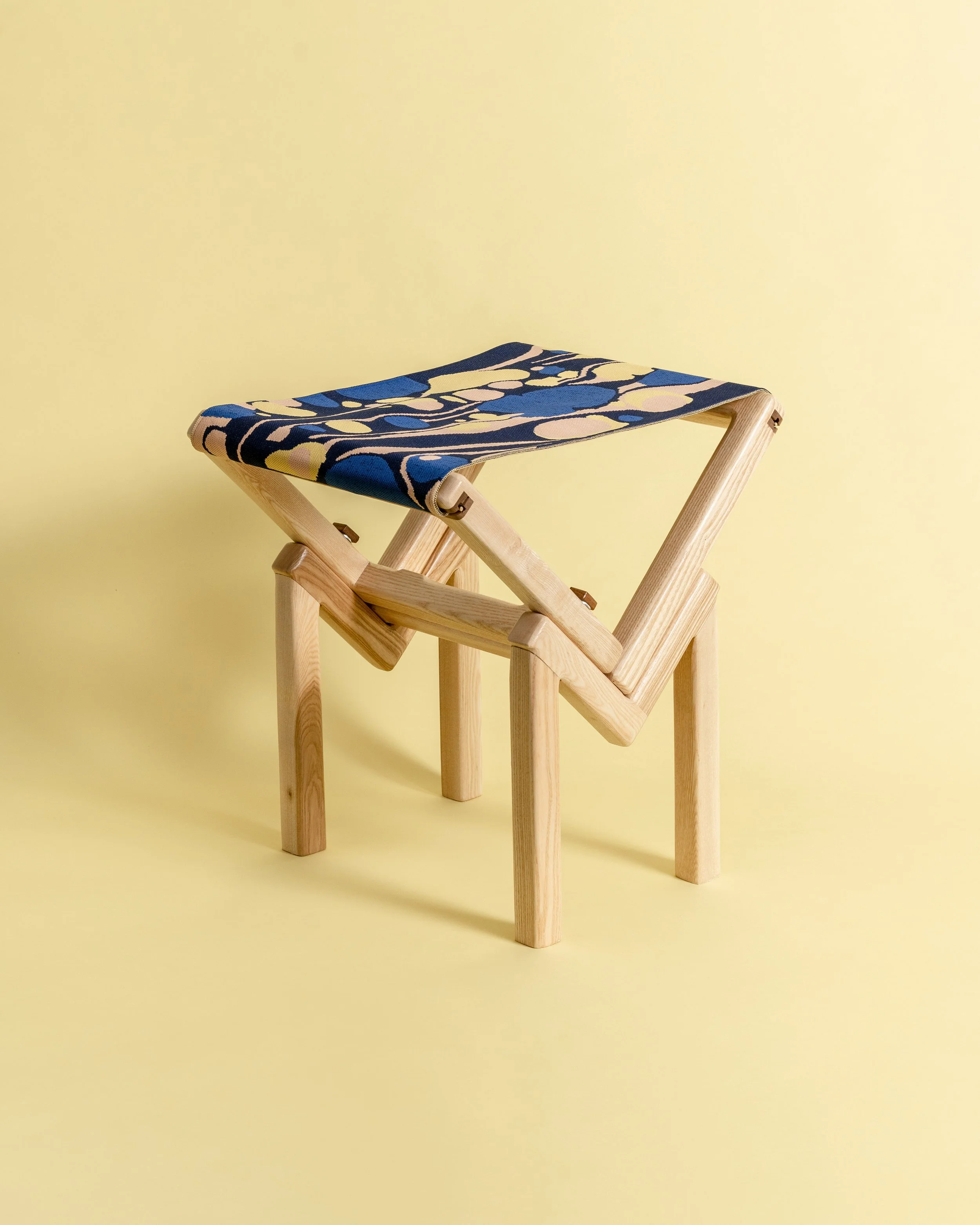 A small wooden stool with a fabric seat, set against a yellow background.