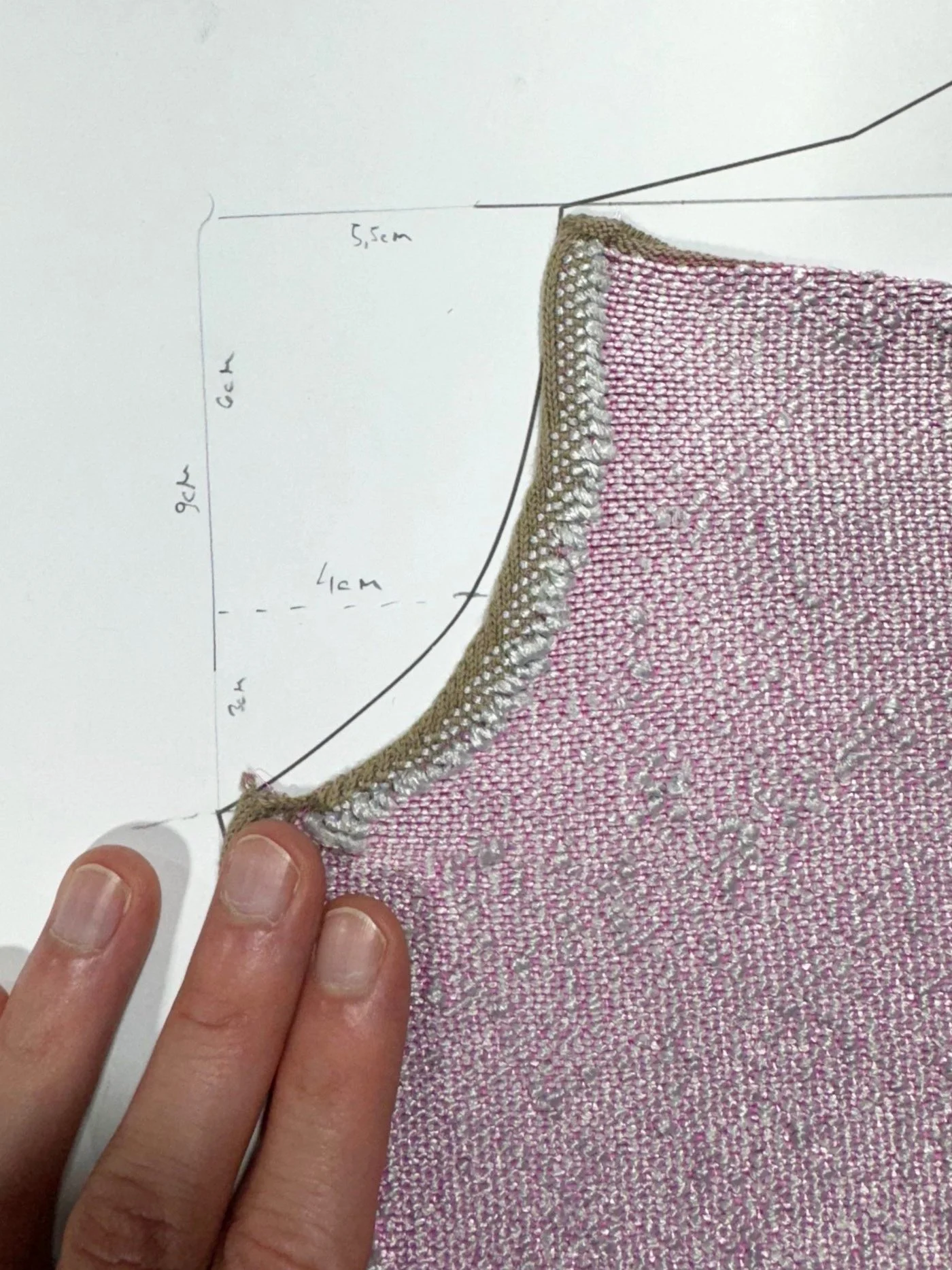 Hand holding pink woven fabric with silver and gold decorative trim, placed on a white surface with handwritten fabric measurements.