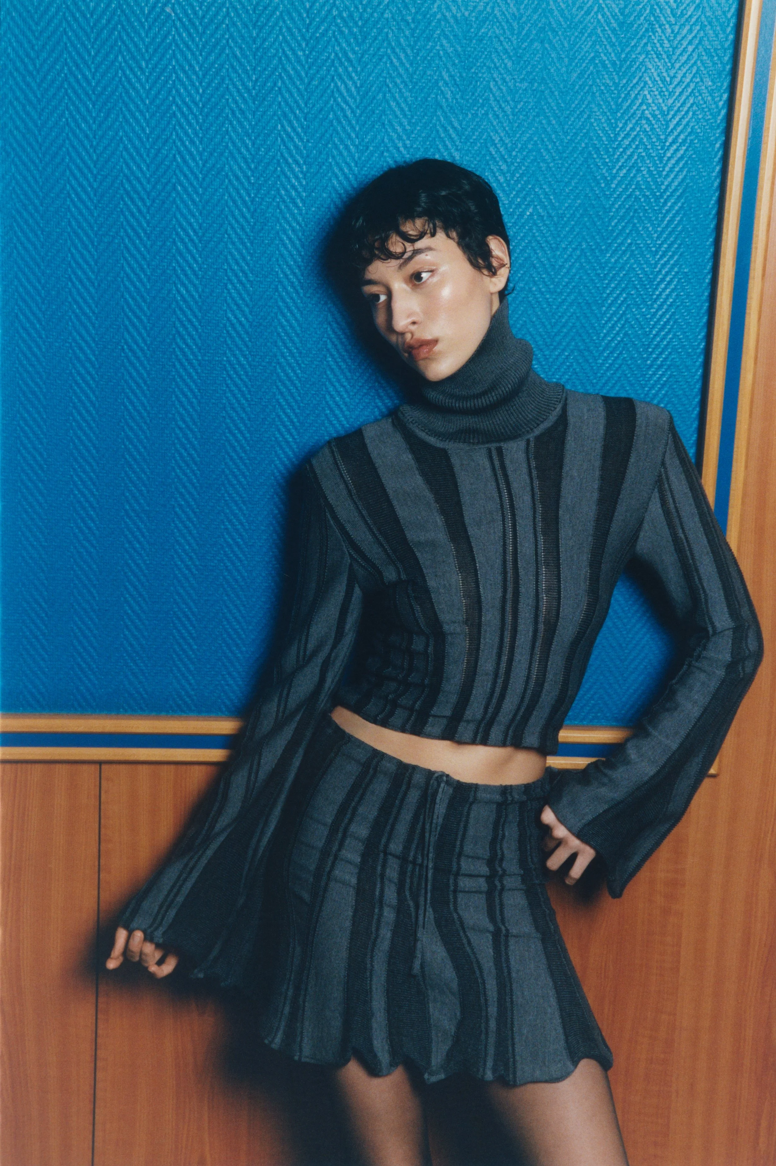 A woman with short curly hair wearing a gray and black striped turtleneck sweater and a matching skirt, standing against a blue wall with wood paneling.