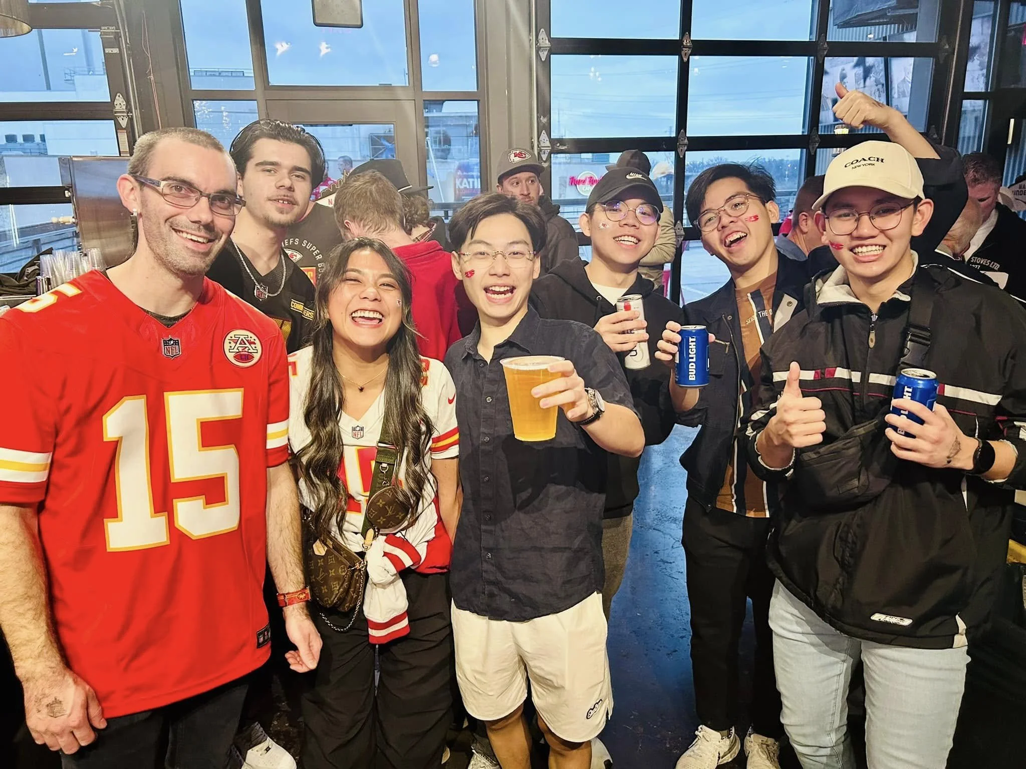 Group of people at a sports bar, many wearing Kansas City Chiefs apparel, smiling and holding drinks, with a large window and cityscape in the background.