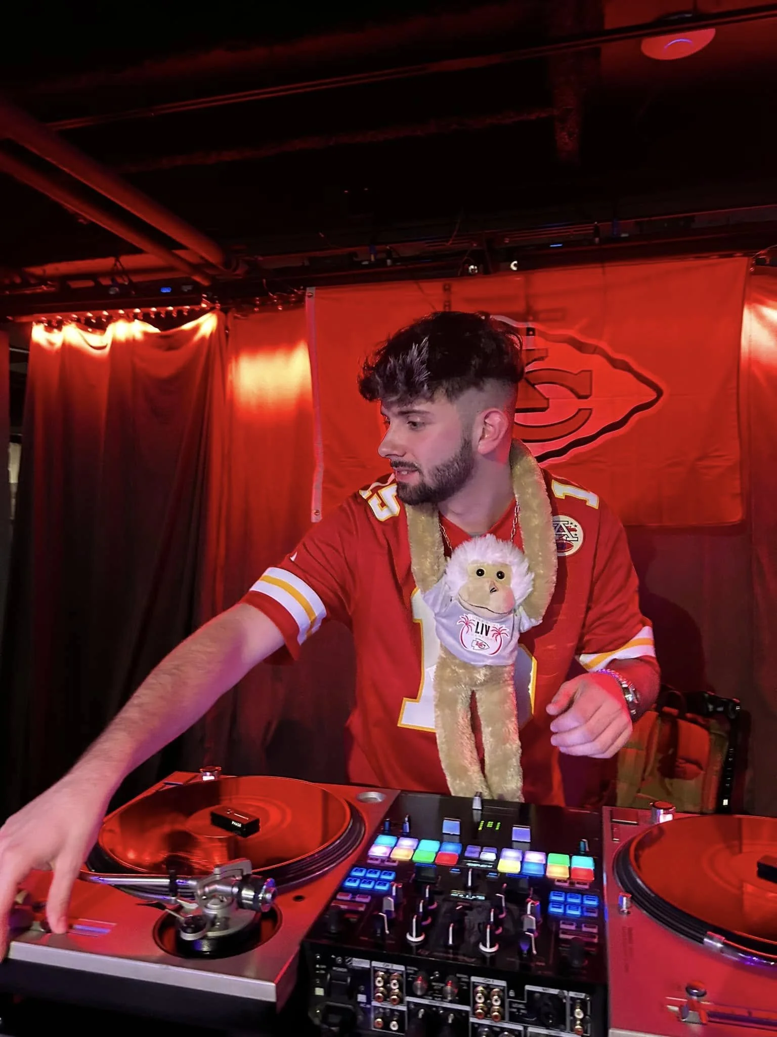 A man wearing a Kansas City Chiefs football jersey and a festive Christmas scarf with a monkey face design is DJing at a party. He is operating a DJ mixer and turntables, with a backdrop featuring the Kansas City Chiefs logo, in a dimly lit room with red lighting and curtains.