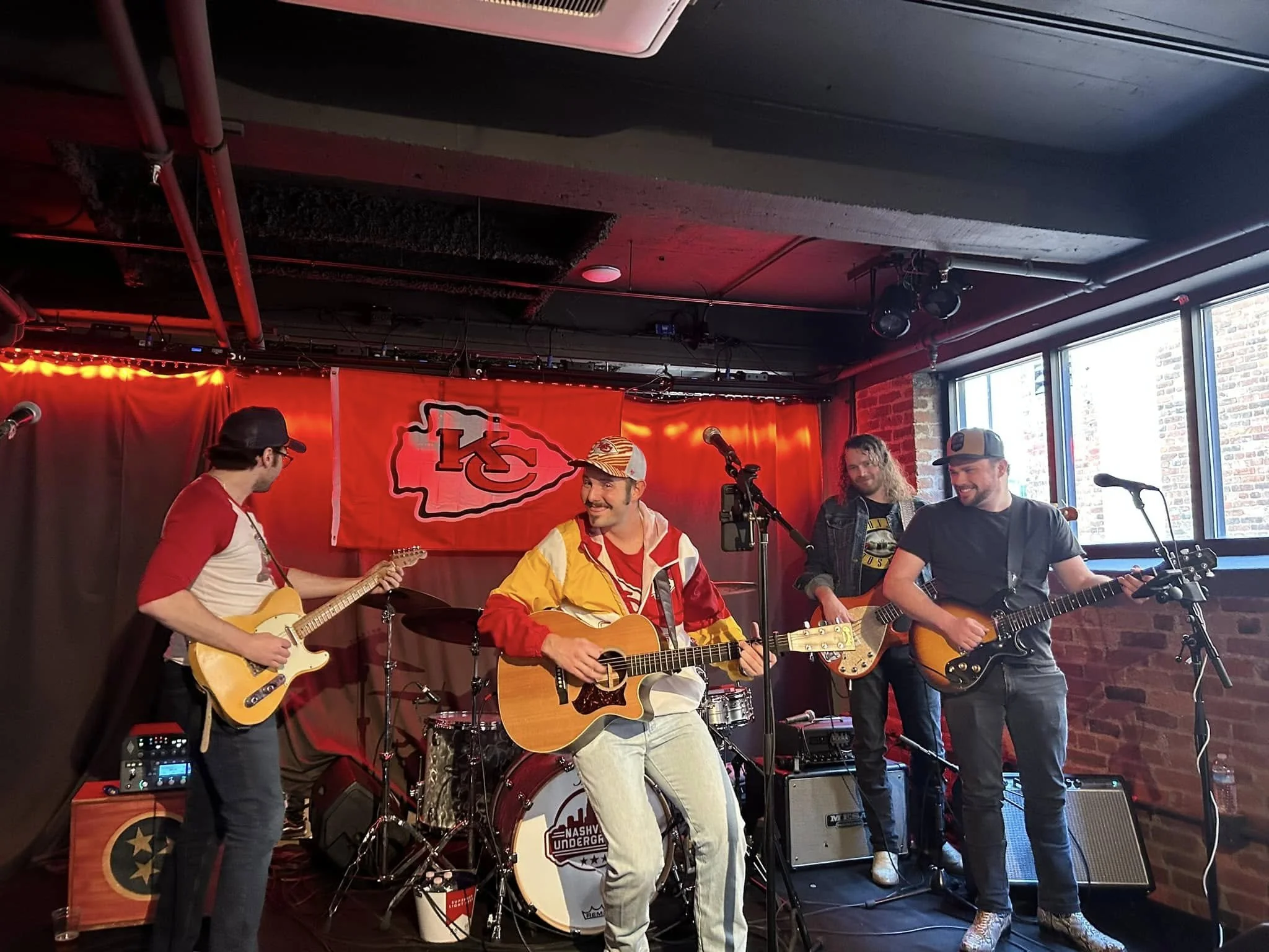 A group of five musicians performing on a stage in a venue with brick walls and large windows. Four of them are playing guitars, and one is sitting behind a drum set. They appear to be enjoying the performance, with smiles and relaxed postures. The background features a red curtain and a Kansas City Chiefs flag.