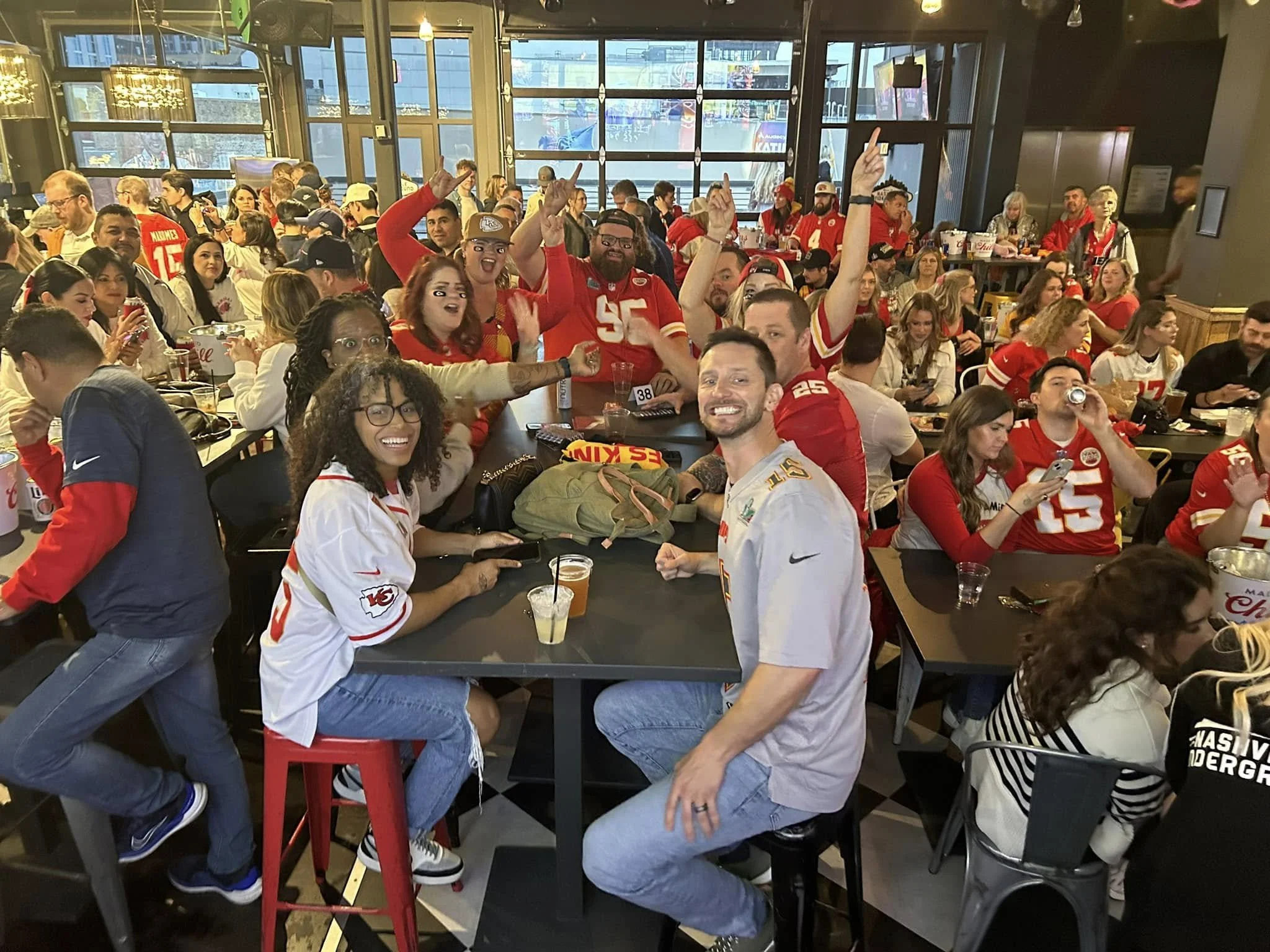 A lively crowd of sports fans gathered inside a bar, most wearing red and white jerseys, celebrating and cheering while watching a game on television screens. Many have their hands raised, and some are drinking beverages.