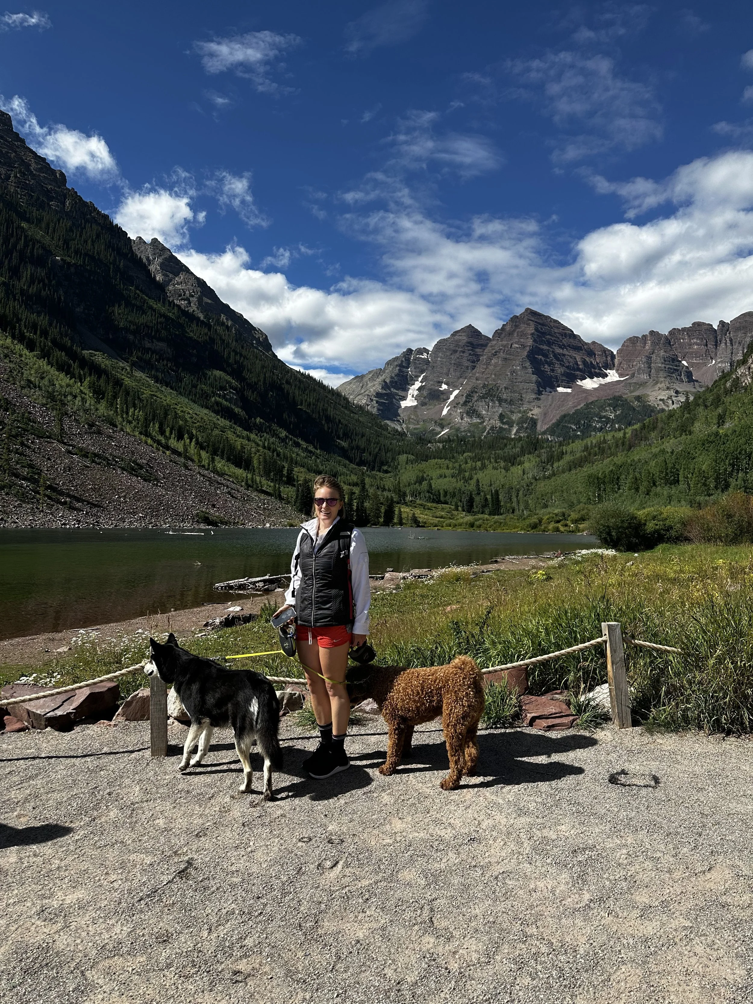 Woman with sunglasses walking two dogs—a black and white husky and a curly brown dog—on a gravel trail near a Colorado mountain lake under blue sky, reflecting connection, grounding, and the healing power of nature in therapy and counseling.