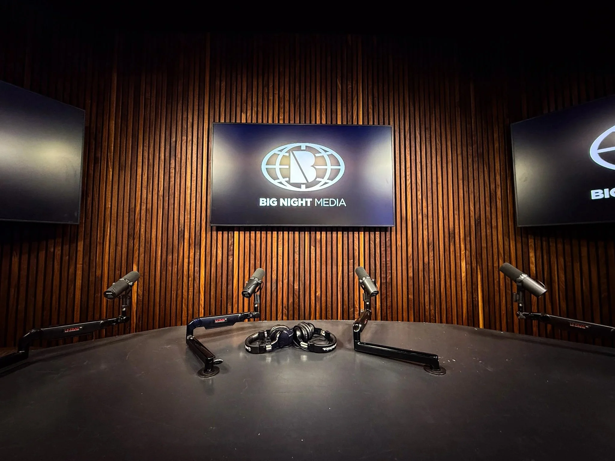 Our podcast studio with a curved black table, microphones, and headphones, against a wooden slat wall with three large screens that can display your podcast logo.