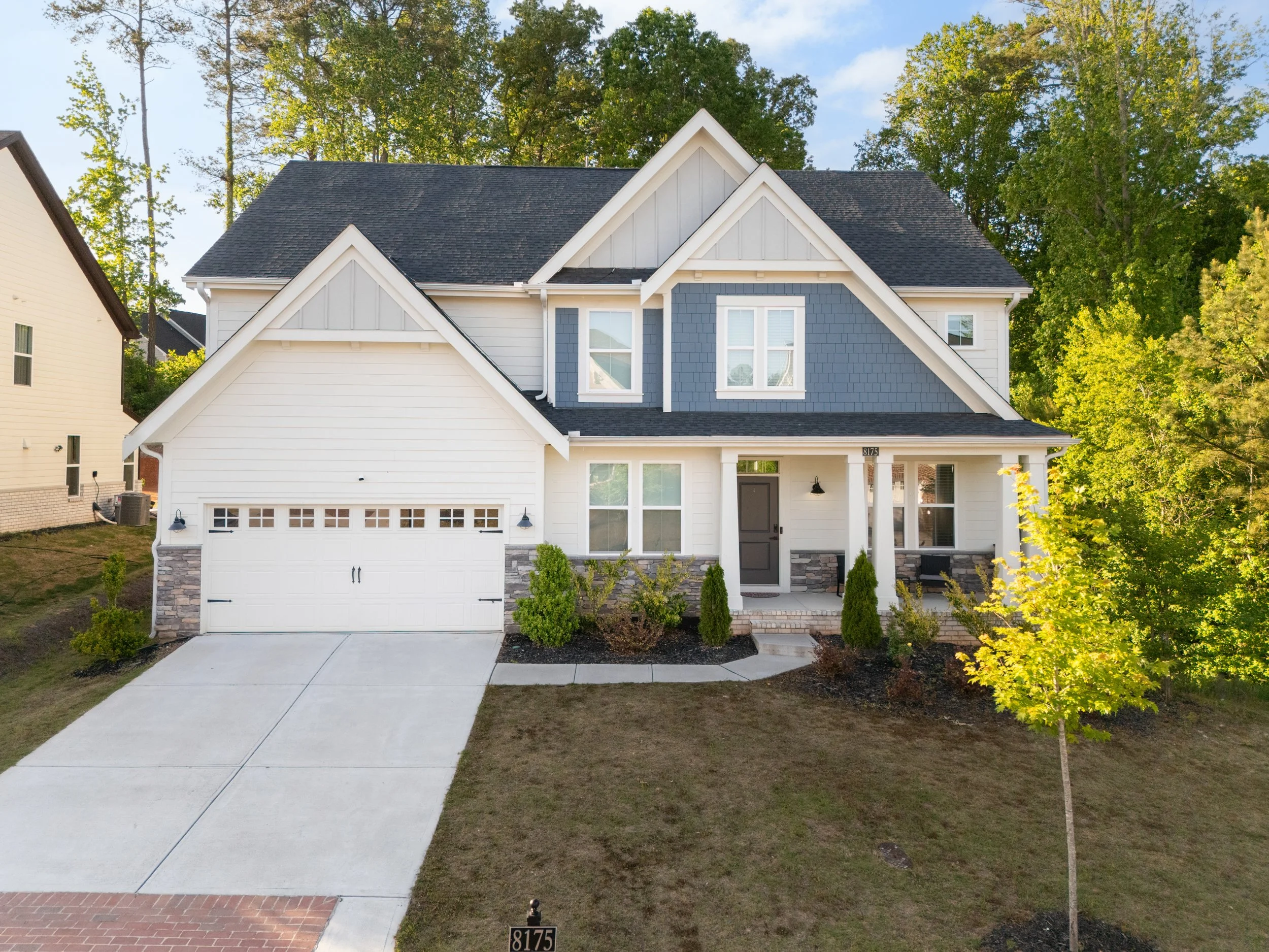 Front exterior of 8175 scenic ridge way in ball ground georgia a two story craftsman style home featuring a white and slate blue board and batten siding, stacked stone accents, a two car garage and covered from porch with georgia trees