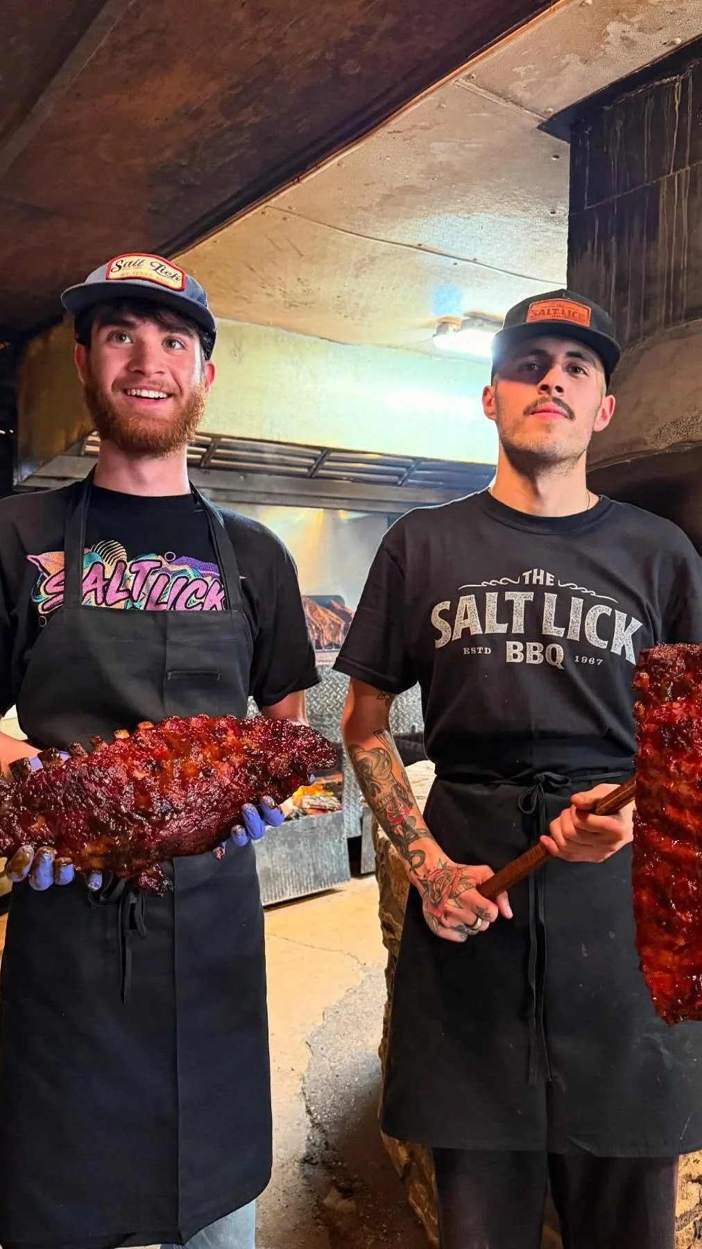 Two men stand in a barbecue restaurant, holding racks of smoked meat. They wear black T-shirts and aprons, and both hats with 'Salt Lick' branding. The man on the left is smiling and holding a large rack of ribs with red glazed sauce, while the man on the right is serious, holding a smaller rack of ribs with a wooden tool. The background features a rustic kitchen setting.
