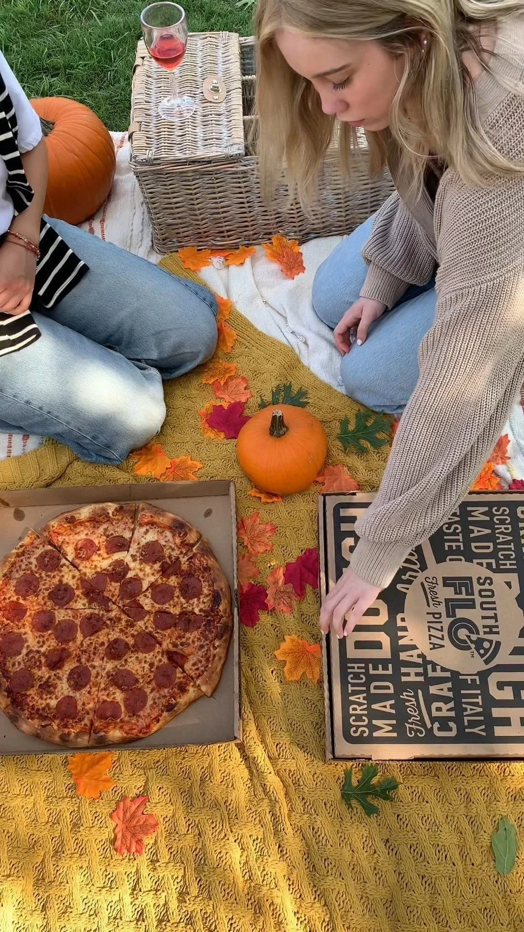 Two women having a picnic outdoors on a yellow blanket with autumn leaves. There is a pumpkin, a pizza box with a pepperoni pizza, a glass of red wine, and a wicker basket on the blanket.