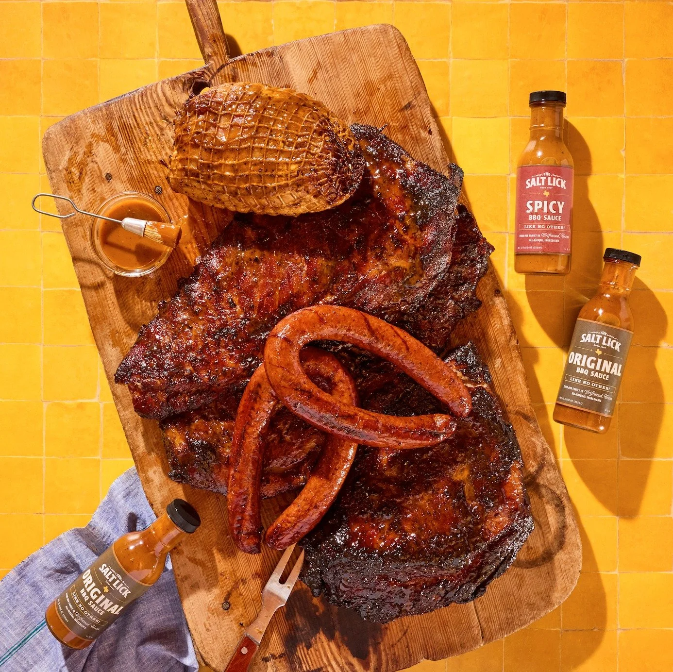 Barbecue meal on a wooden board with ribs, sausage, and a baked potato, surrounded by bottles of BBQ sauce and a small bowl of barbecue sauce.