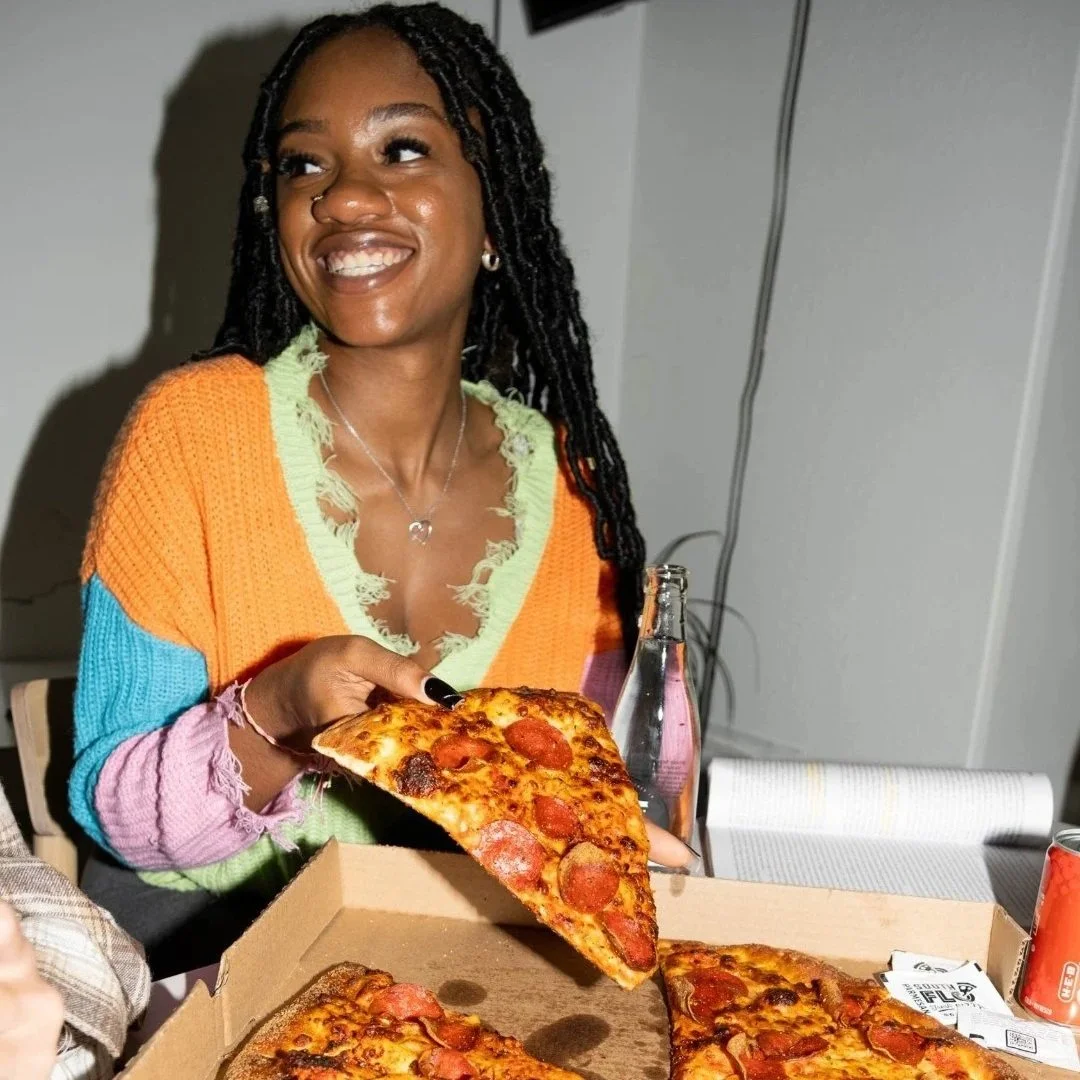 A woman with long black braids, smiling and holding a slice of pepperoni pizza, sitting at a table with a pizza box, a bottle, and a can.