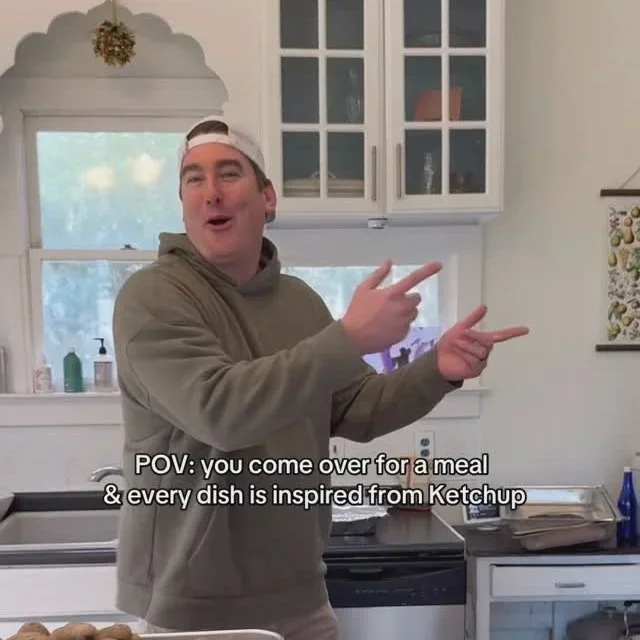 Young man wearing a backward white cap and a hoodie, pointing with both hands in a kitchen. Subtitles say: "POV: you come over for a meal & every dish is inspired from Ketchup."