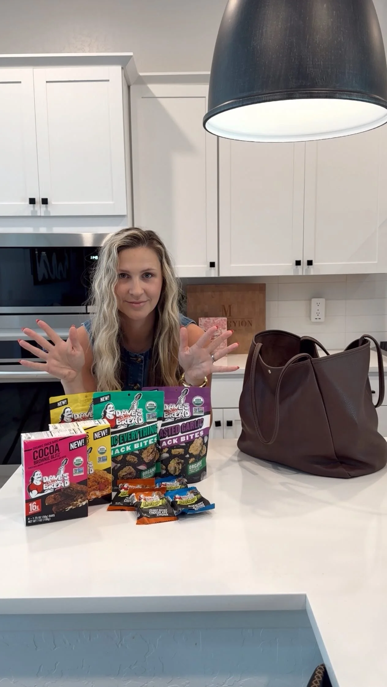 Woman sitting at a kitchen counter with various snack products in front of her, making a gesture with her hands, with a brown purse on the counter to her right.