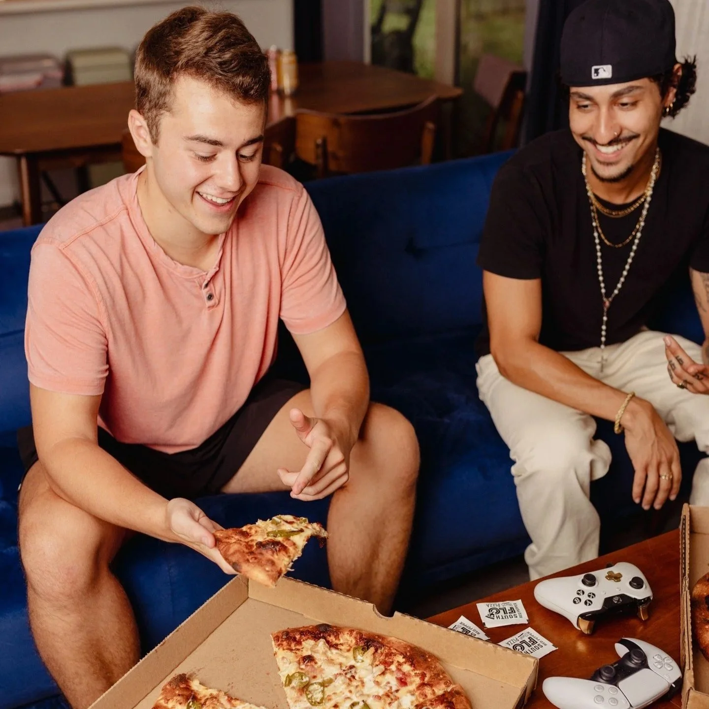 Two young men sitting on a blue couch, sharing a pizza. One is holding a slice of pizza and smiling, while the other, wearing a black cap and jewelry, is also smiling. There are gaming controllers and pizza boxes on the table in front of them.