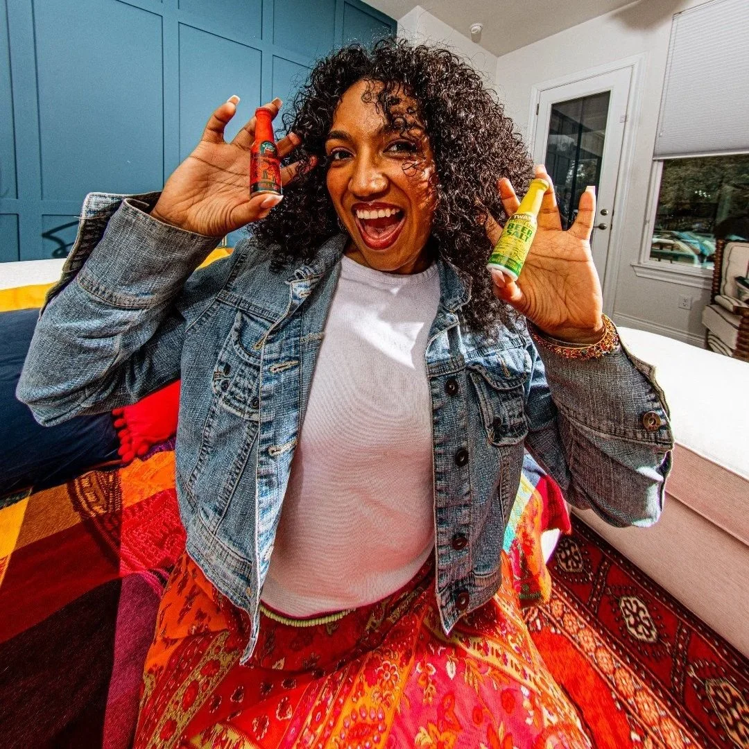 A woman with curly hair smiling and holding two bottles of hot sauce, one in each hand, indoors with a colorful patterned rug and a blue wall in the background.
