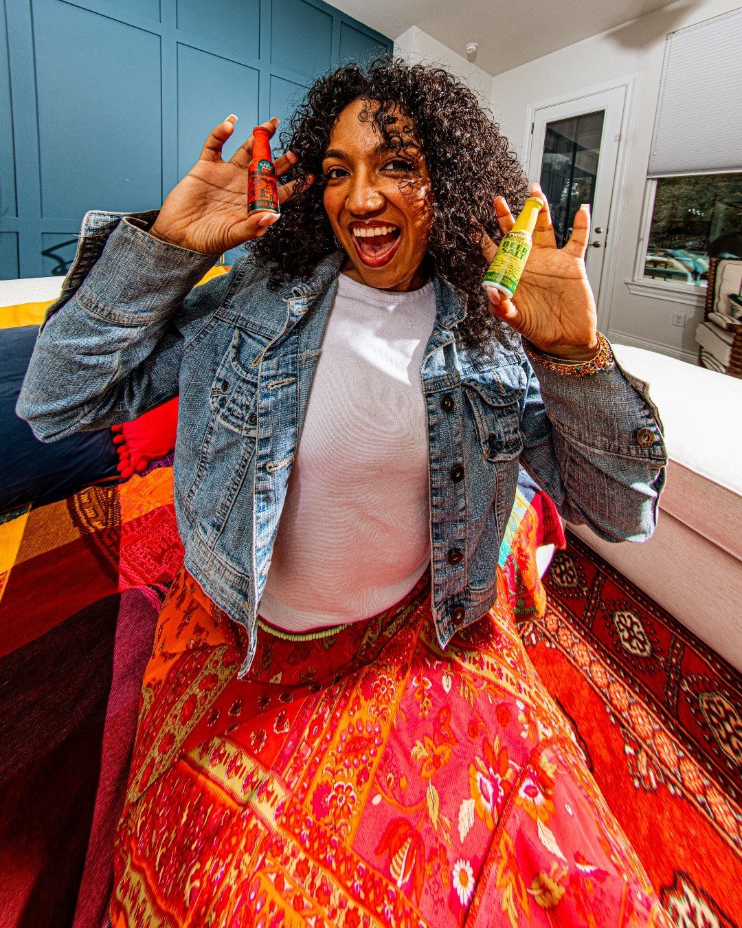 A woman with curly hair smiling and holding up two small bottles, one red and one green, in a colorful indoor setting.