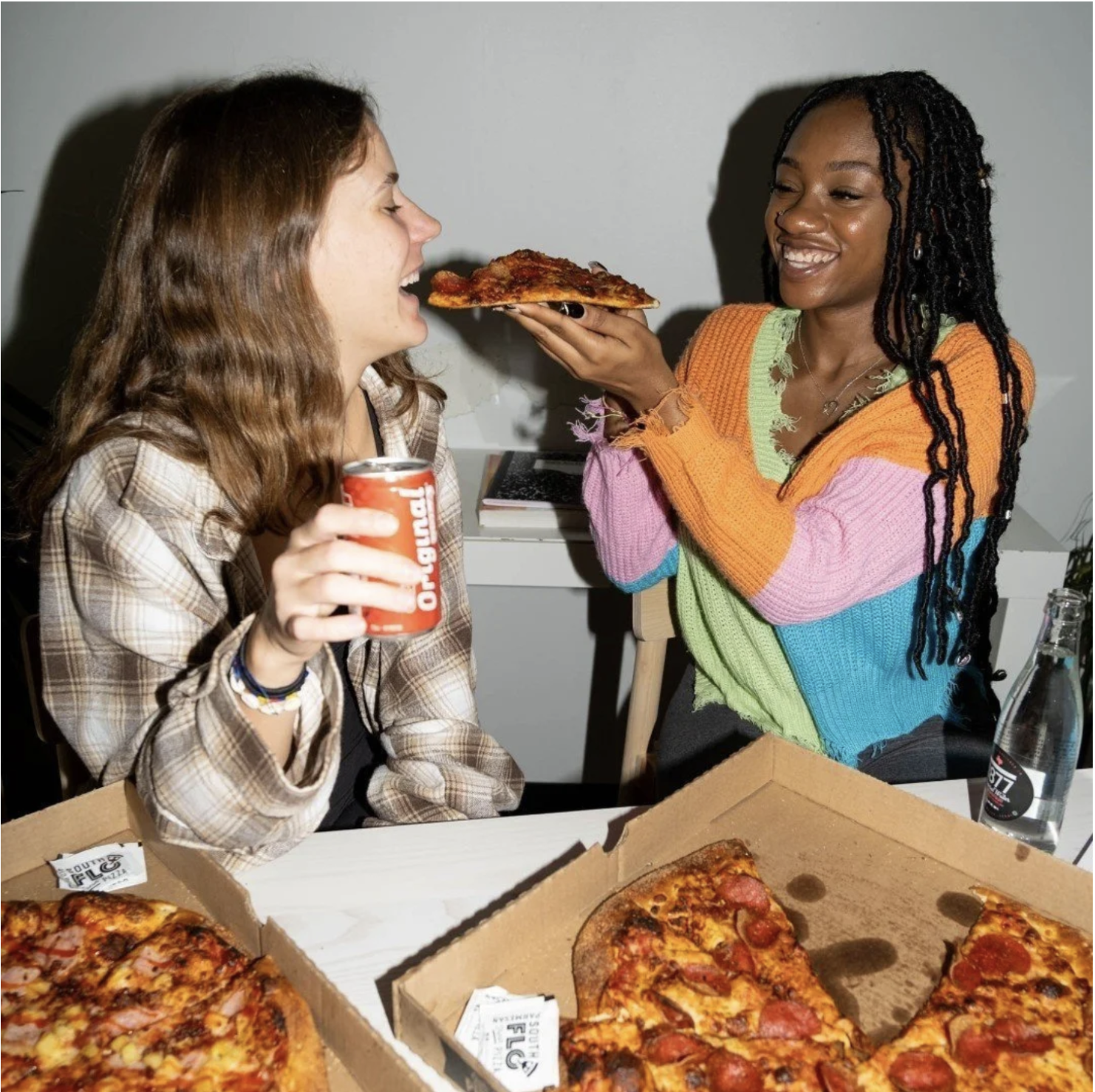 Two women sharing a pizza, with one woman feeding a slice to the other. The woman on the left has brown hair and is holding a red soda can, while the woman on the right, with black braided hair, is wearing a multicolored sweater. There are open pizza boxes with pizza slices on the table.