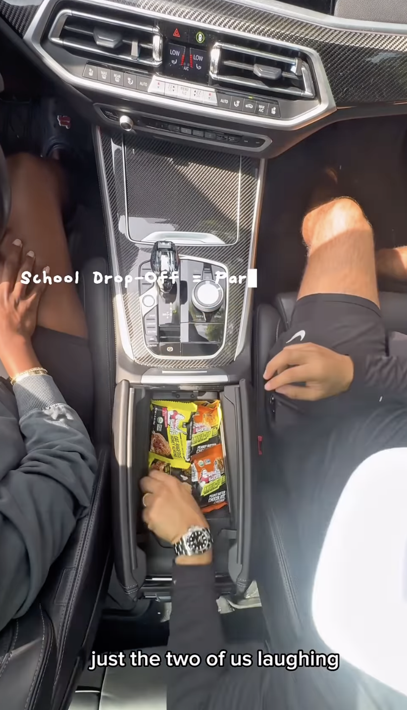 Inside a car, two people are sitting in the front seats. The center console contains a compartment filled with snack bars. One person is reaching into the snack compartment. The dashboard controls are visible at the top of the image. The caption indicates this is during a school drop-off and they are laughing.