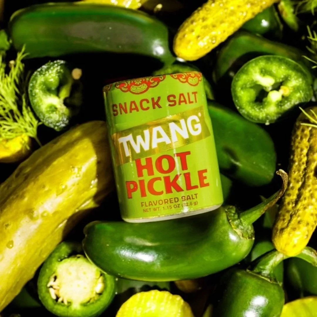 Can of Twang Hot Pickle flavored salt surrounded by cucumbers, jalapeños, and yellow mini corn cobs.