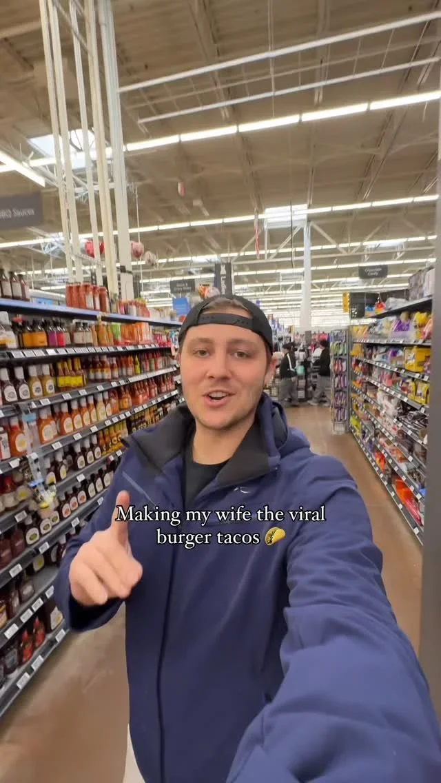 A man taking a selfie in a grocery store aisle, wearing a navy jacket and black cap backwards, with a caption 'Making my wife the viral burger tacos' and a taco emoji.