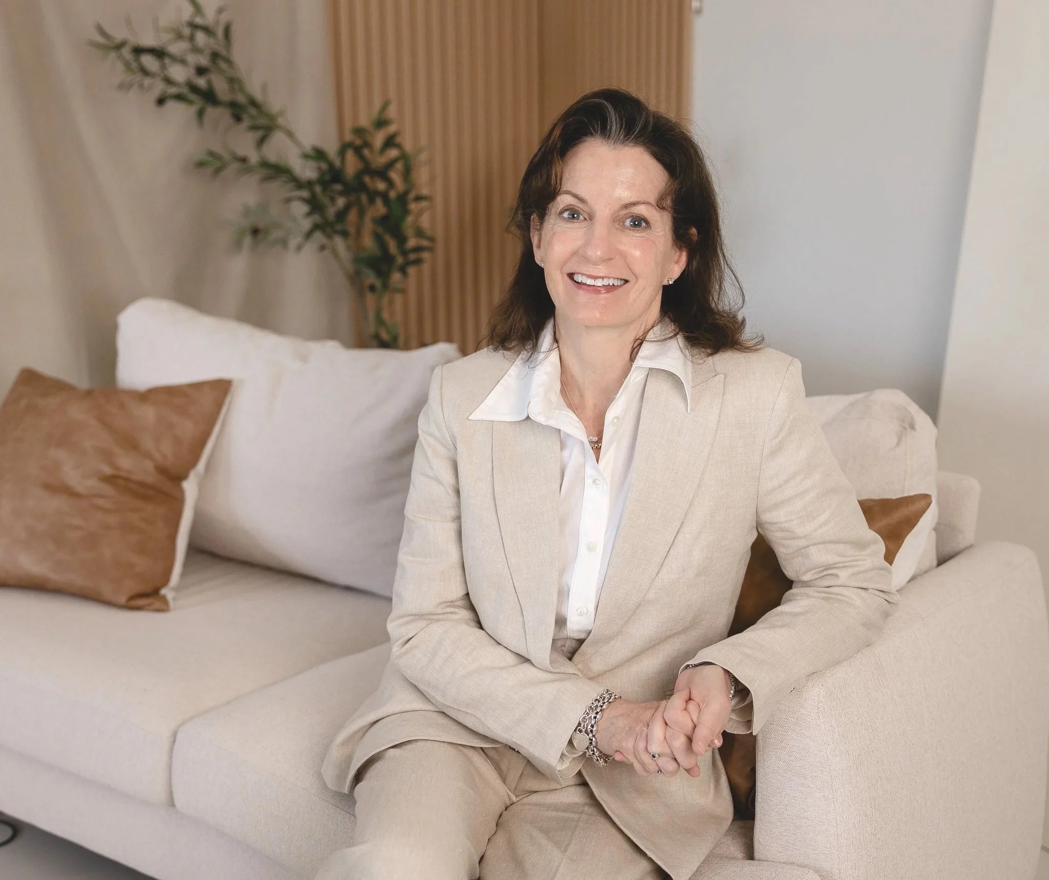 A woman sitting on a beige sofa, smiling at the camera, wearing a light-colored suit.