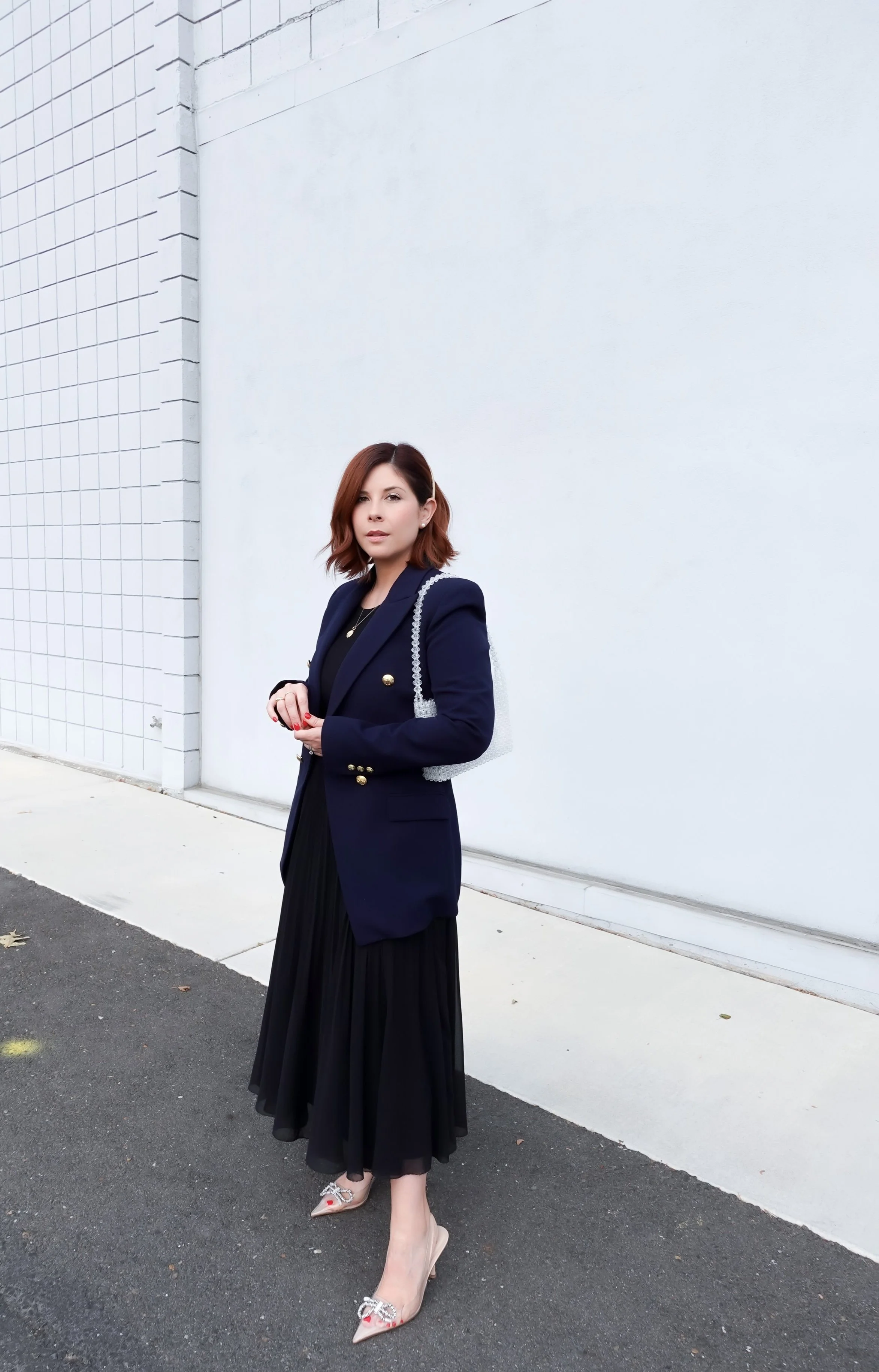 A woman with shoulder-length brown hair wearing a navy blazer, black pleated skirt, and beige high heels standing on a sidewalk against a white wall.