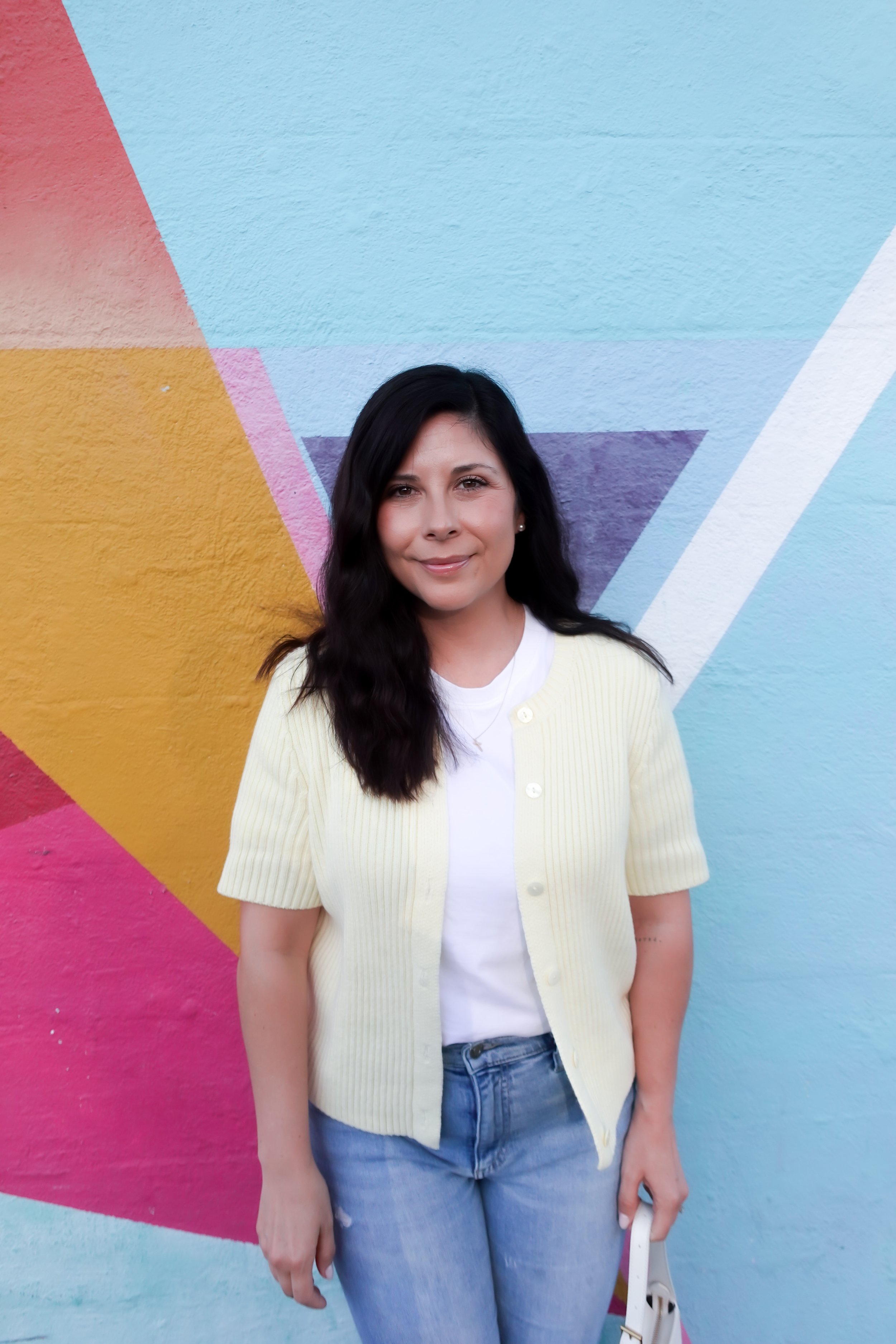 A woman poses smiling in front of a vibrant geometric street art mural. She wears a short-sleeve pale yellow cardigan over a white t-shirt, light-wash blue jeans, and clear heels with bow details. She is carrying a small white shoulder bag.