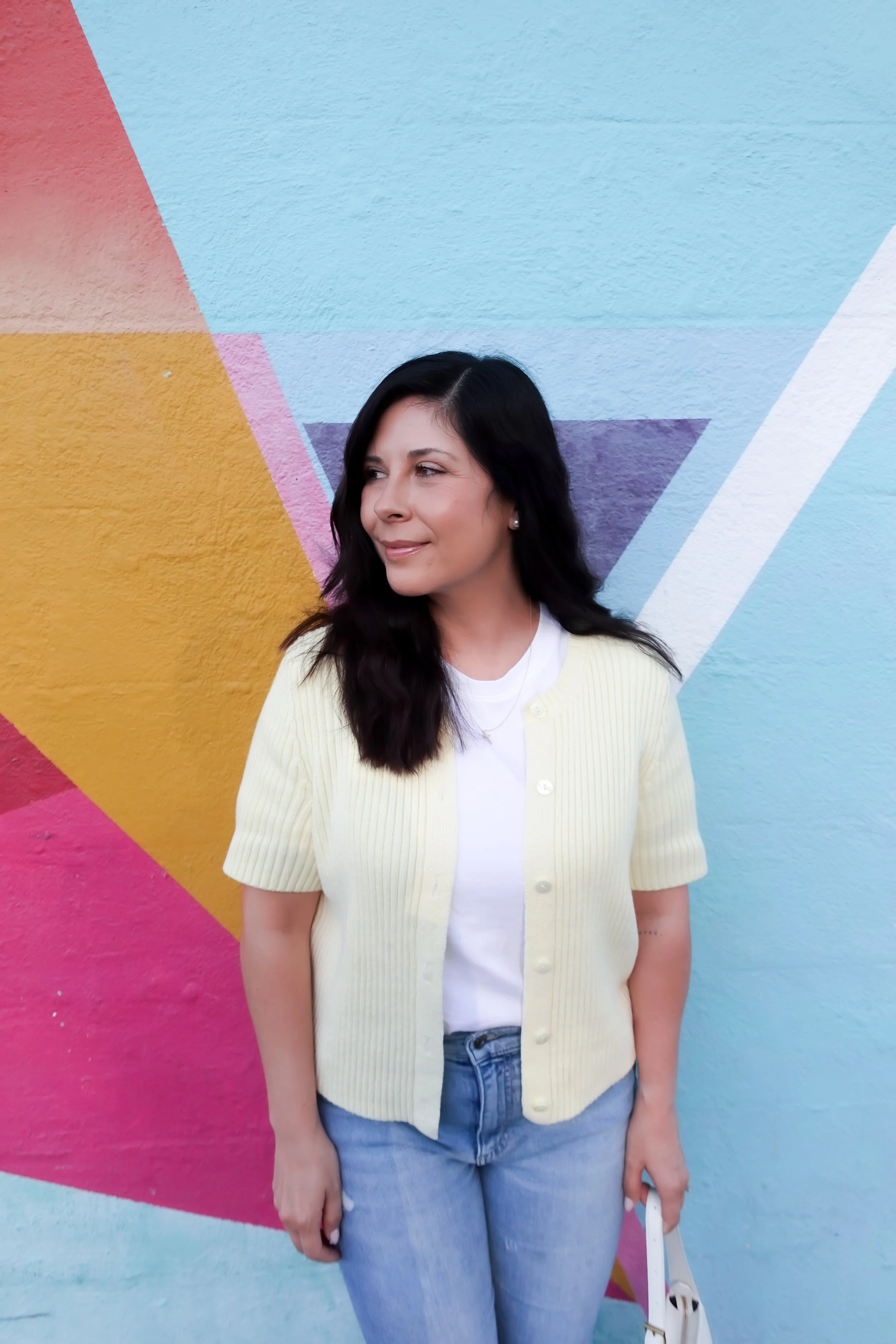 A woman poses smiling in front of a vibrant geometric street art mural. She wears a short-sleeve pale yellow cardigan over a white t-shirt, light-wash blue jeans, and clear heels with bow details. She is carrying a small white shoulder bag.